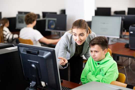 Caring friendly female teacher calming upset tween schoolboy sitting at computer in classroom, helping him complete assignment during lesson