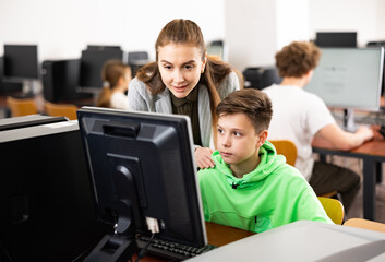 Fototapeta premium Positive female teacher helping focused preteen schoolboy studying in college library computer lab..