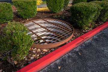 Large round stormwater containment drain with rusty iron grate in a median garden, red fire lane...