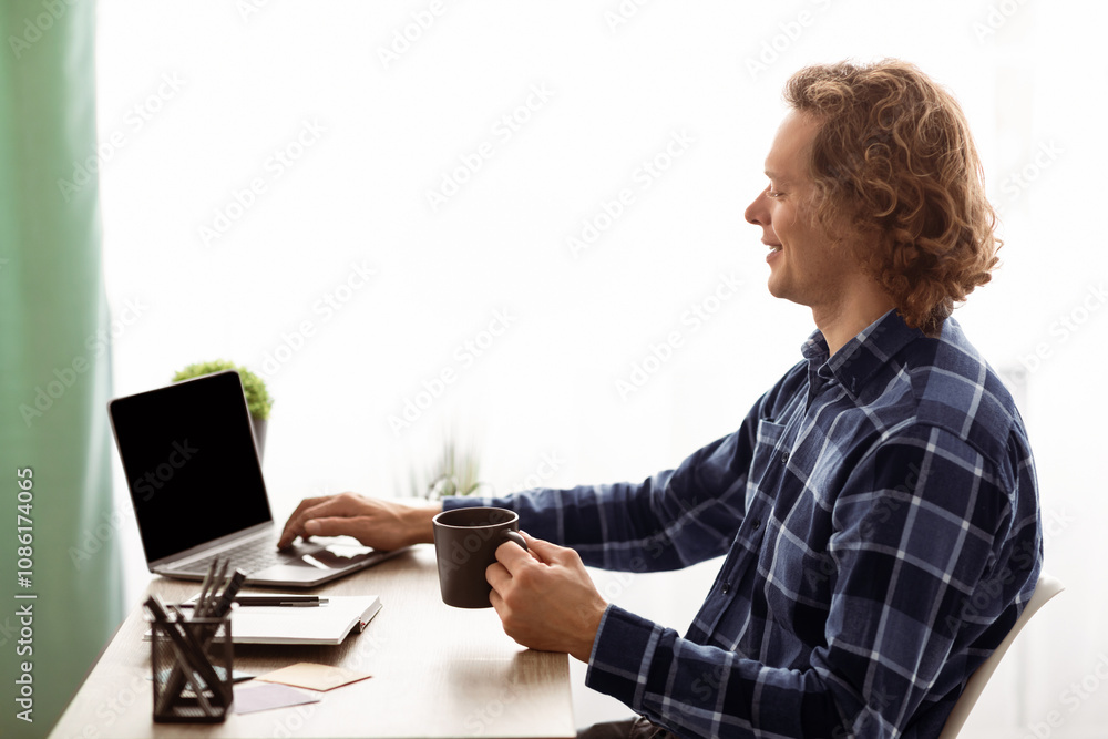 Poster side view of millennial guy using laptop with empty screen drinking coffee sitting at workplace in m