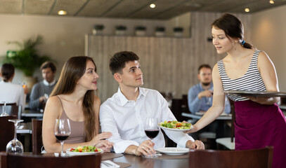 Young woman waitress brings order from couple young man and young woman in restaurant