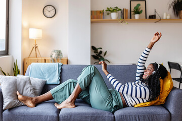 Young happy black woman dancing and enjoying online music while lying on comfortable sofa enjoying day off at home.
