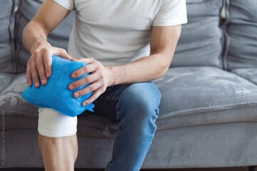 Close-up of a man sitting on a couch, holding a blue ice pack on his bandaged knee, highlighting injury care at home.