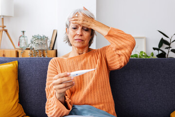Senior woman is sitting on her sofa, holding a thermometer and feeling sick with a fever