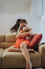 Obraz premium A young girl in a pink dress hugs her stuffed toy while sitting comfortably on a sofa, creating a warm and cozy atmosphere.