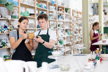 Young woman teacher in apron examines ceramic cup made by teenage boy student
