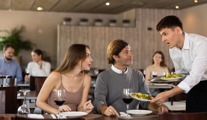 Male waiter serving fresh vegetable salads to married couple guests visiting restaurant for dinner, sitting at table with glasses of red wine