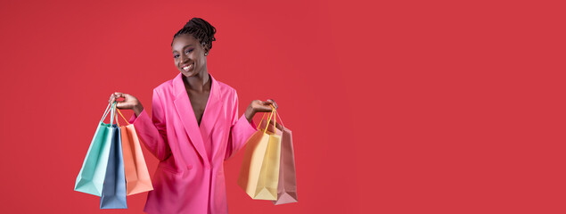 Fototapeta premium Happy young African American woman holding colorful shopping bags in her hands, posing in pink costume on red background, panorama with copy space