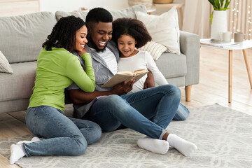 Happy african american family reading book together, staying at home during coronavirus quarantine, sitting on floor in living room, bonding