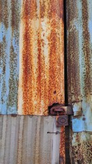 Close-up of a rusty, weathered, corrugated metal sheet door locked with a rusty padlock, showcasing peeling paint and significant signs of corrosion over time.