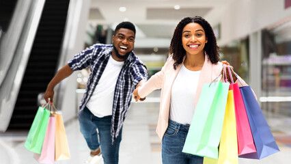 Sales Season. Excited Black Woman Pulling Boyfriend To Shopping Store To Buy New Clothes, Fooling And Running In Mall Together