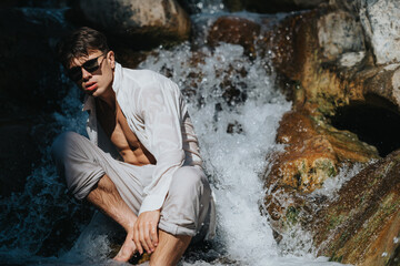 Young man in sunglasses leaning against rocks in a waterfall, wearing a wet white shirt. A fashionable and adventurous outdoor scene.