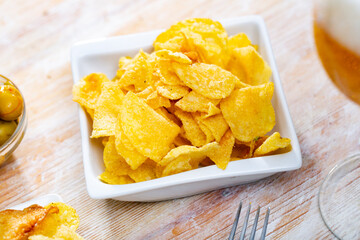 There is portion of yellow crispy potato chips on small plate. Popular snack is complemented by bowl with delicate green olives