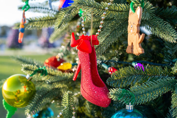 A bright red high-heel stiletto with a red bow. The sultry ruby slipper is covered with glitter. The Christmas decoration hangs on a real fir tree with glass balls, dog ornament, and mini lights. 