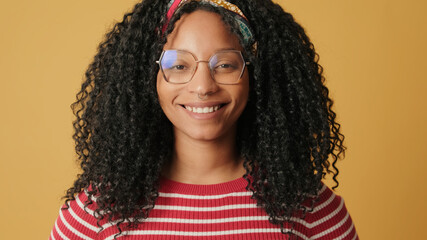 Close up, young woman with curly hair wearing glasses wearing striped sweater, using mobile phone, on yellow background in the studio