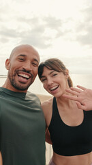 A multi-ethnic man and woman takes a photo of themselves, greeting followers and laughing, takes a selfie for content. Male and female athletes by the sea using phone camera