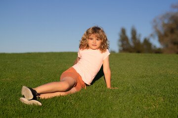 Fototapeta premium Outdoor portrait of cute kid wearing sunglasses, summer hat, t-shit and short. Happy children rest fun outdoors. Kids relaxing in spring park. Boy sitting on green grass.