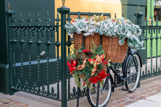 A black two-wheel bicycle chained to a dark green wrought iron fence. The butcher's bike has a large wicker basket filled with greenery and Christmas flowers. There's a wreath with red bows on front. - Powered by Adobe