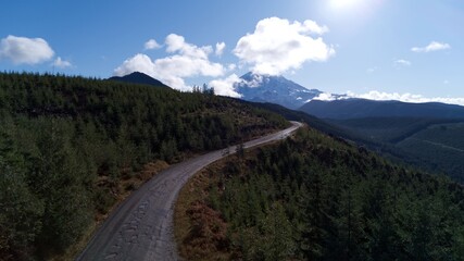 A scenic aerial view of a winding road surrounded by lush forested hills leading toward a majestic Mount Rainier under a blue sky with scattered clouds, capturing the beauty of the Pacific Northwest.
