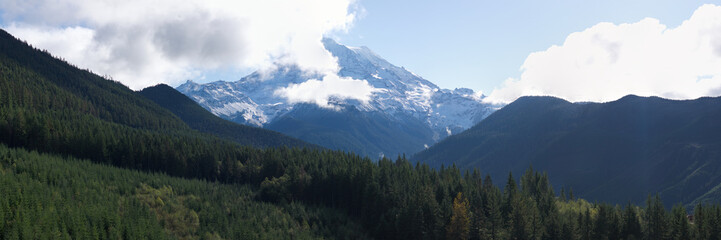 A scenic panorama capturing Mount Rainier partially covered by clouds, surrounded by lush, forested hills under a bright sky.