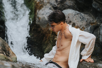 Young man wearing a white shirt and sunglasses, enjoying nature by a waterfall. Relaxation, adventure, and natural beauty.