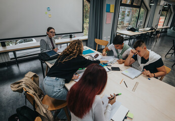 A diverse group of students collaborating in a modern classroom setting. They are engaged in a study session, discussing and taking notes with a teacher overseeing their work.
