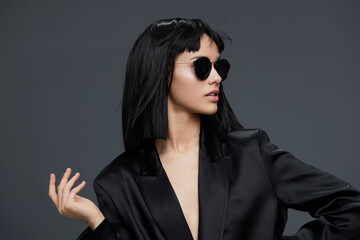 Beautiful asian woman in a black suit and sunglasses confidently posing against a gray background,...