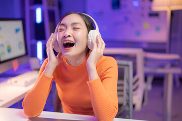 Young asia businesswoman wearing headphones using desktop computer listening to music and singing while working late night at home office