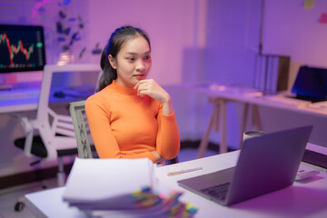 Young asian businesswoman in orange sweater works late at modern office desk, focused on laptop with stock market charts in background