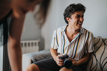 Joyful young man holding a camera while sitting on a couch, wearing a striped shirt in a relaxed...