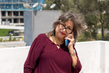 Senior Latina woman with glasses talking on her cell phone on a terrace with trees and buildings...