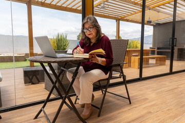 Senior businesswoman writing with a pen in a notebook next to a laptop on a terrace pergola in the morning