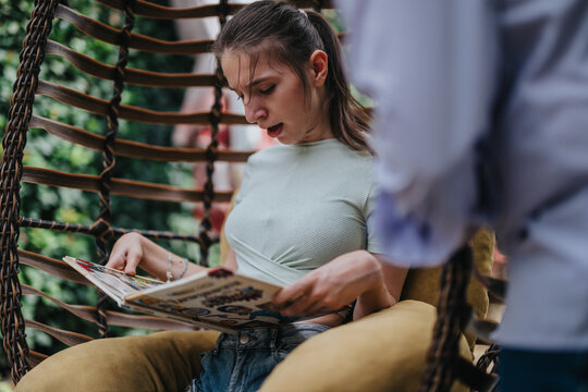 A young girl with a disability is engrossed in reading a book while sitting comfortably in a woven swing chair, surrounded by greenery.