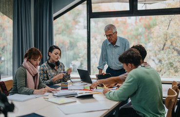 A group of university students working together on a project while their senior professor provides guidance and assistance in a classroom with large windows and natural light.