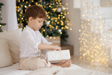 A young boy sits on a bed with a white blanket and a white shirt, opening a present. The room is decorated with Christmas lights and a Christmas tree. The boy appears to be excited about his gift.