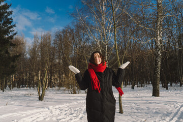 A smiling woman with a red scarf enjoys a snowy winter day in a serene forest setting beneath a bright blue sky. Christmas and New Year celebration concept.