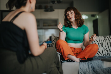 Two young women engage in an animated conversation while sitting on a sofa in a casual, cozy living room. The atmosphere is relaxed and friendly, highlighting social interaction and communication.