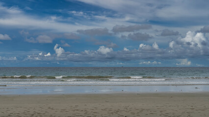 The waves of the endless turquoise ocean foam and spread over the beach. Reflection on wet sand. Sand bubbler crabs burrows are visible. A tiny silhouette of a ship on the horizon. Blue sky, clouds.