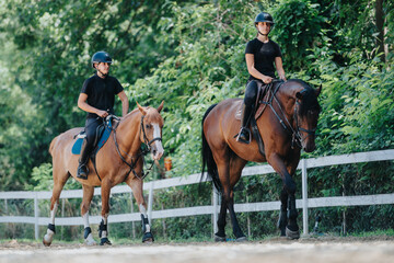 Obraz premium Two horseback riders in black attire navigate their horses through a lush, green forest on a sunny day, enjoying the outdoor ride