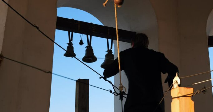Bell ringer skillfully pulling ropes high in Orthodox bell tower at sunset, filling sky with powerful sound. Bells call faithful to prayer, reflecting deep roots of Orthodox spirituality.