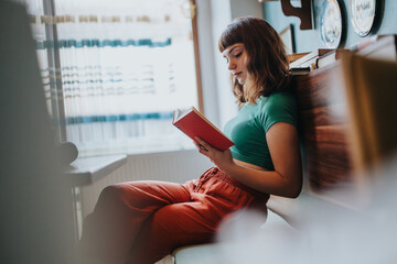 A young woman sits comfortably indoors, engrossed in reading a book, enjoying a serene and calm...