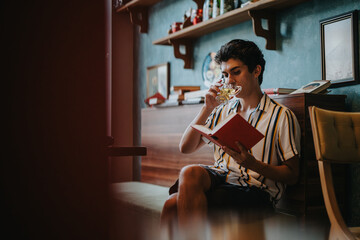 A relaxed young man sips a beverage while sitting and reading a book in a cozy home setting. The...