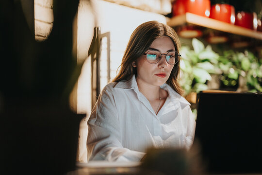 Businesswoman wearing glasses concentrating on her laptop in a modern office setting with lush green plants and vibrant decor.