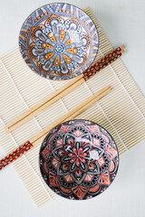 Two empty multi-colored unique patterned china bowls with chopsticks on top and sushi rolls on the base
