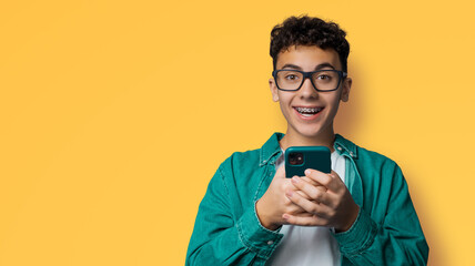 Excited surprised shocked astonished happy curly haired funny young man wear braces, tooth brackets, eyewear glasses open mouth hold typing cell phone cellular smartphone, isolated yellow background