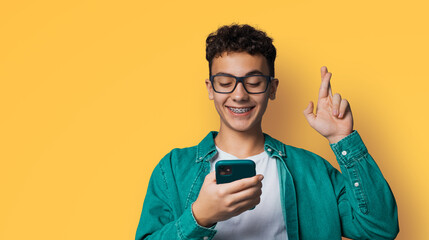 Smiling young man wear braces, tooth brackets, eyewear glasses hold typing text look read cell phone cellular smartphone cellphone, crossing fingers, pray hope for good luck, isolate yellow background