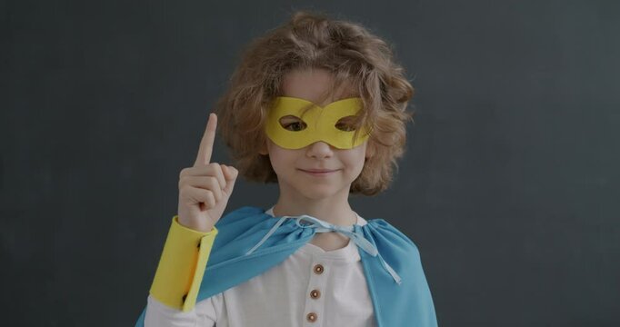Portrait of smart little boy in superhero costume thinking then raising finger having brilliant idea on grey background. Genius kid and emotion concept.