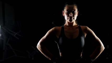 The silhouette of a gorgeous muscular woman in a sports top on a black background, the shadow highlights her muscular body. A bodybuilder poses for the camera in a dark gym.