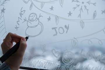 Sustainable responsible holiday decor. Female hand painting snowman painted on window glass indoor...