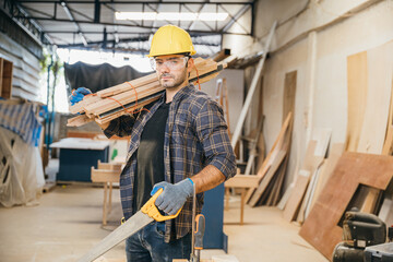 Smiling construction worker in a yellow hardhat and plaid shirt holding wood planks on his shoulder and holding saw. Ideal for carpentry, woodworking in furniture workshop, National Carpenters Day